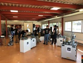 Robotino Roboter mit Stationen im Hintergrund und Studierende im Austausch