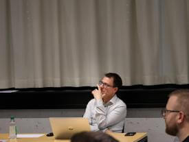 Eine Person sitzt am Tisch mit Laptop und Wasserflasche und beobachtet das Geschehen.